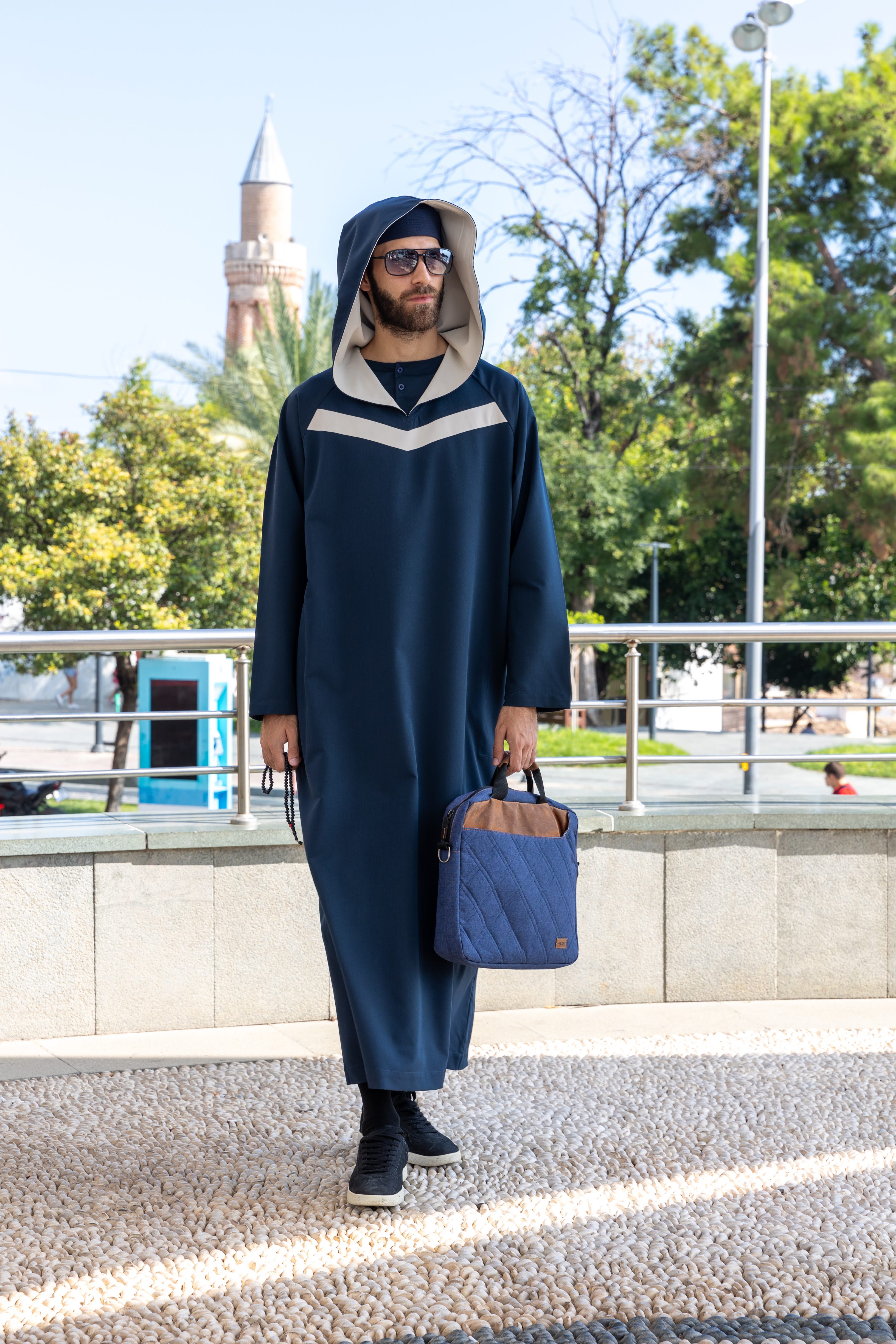 Man in a blue hooded robe holding glasses and a black bag against a white background.