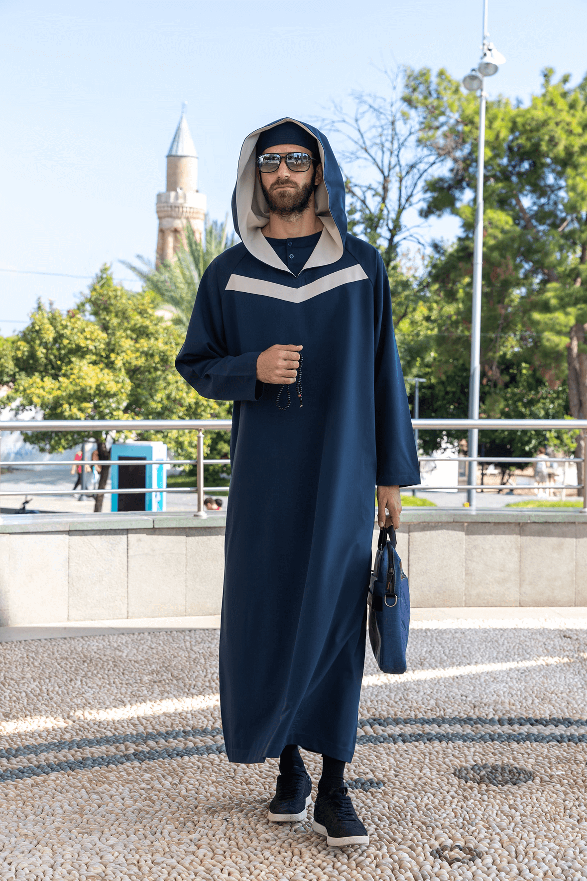 Man in a blue hooded robe holding glasses and a black bag against a white background.