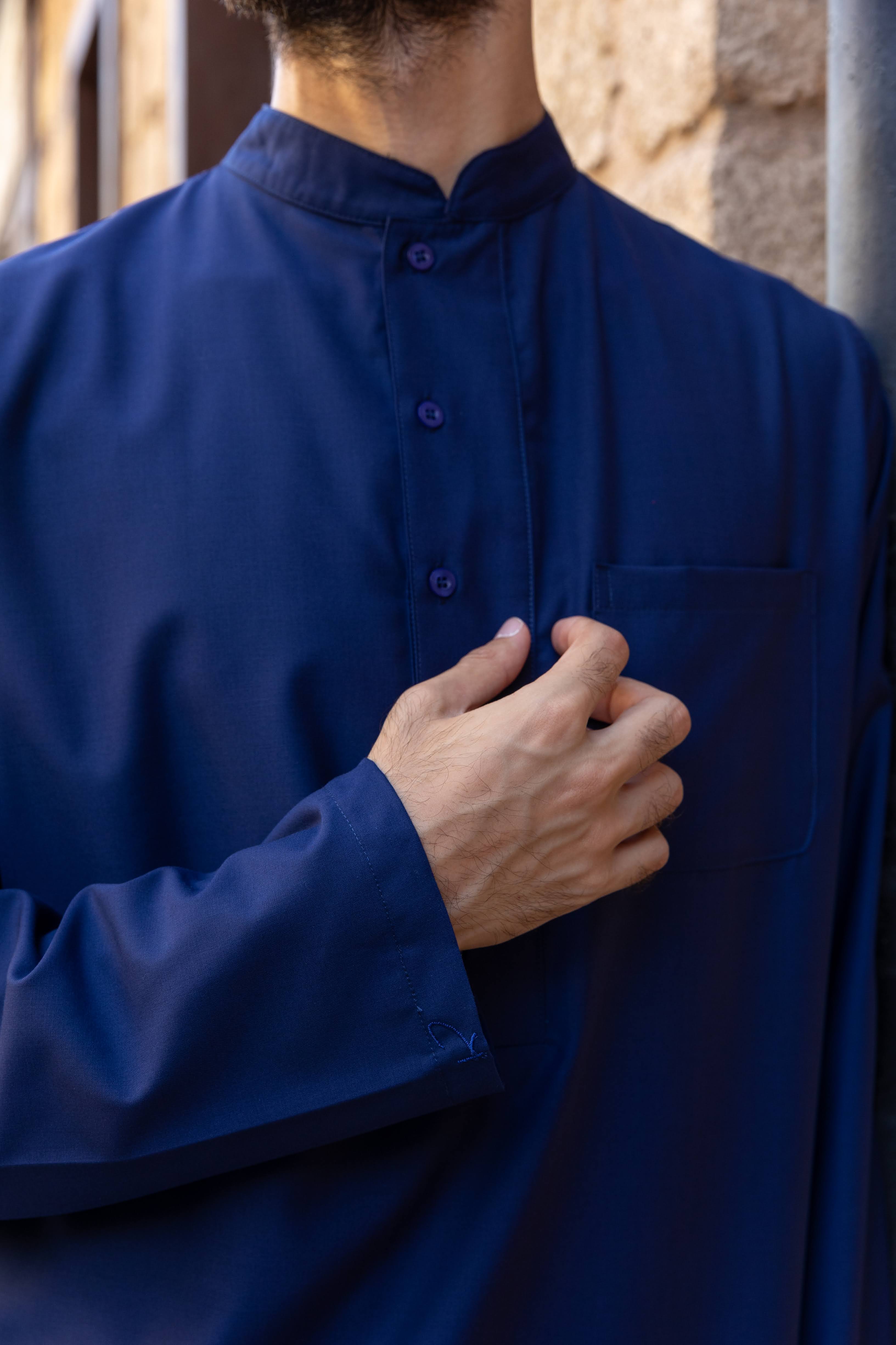 A person in a dark hooded robe, holding a string of beads, standing with back turned.