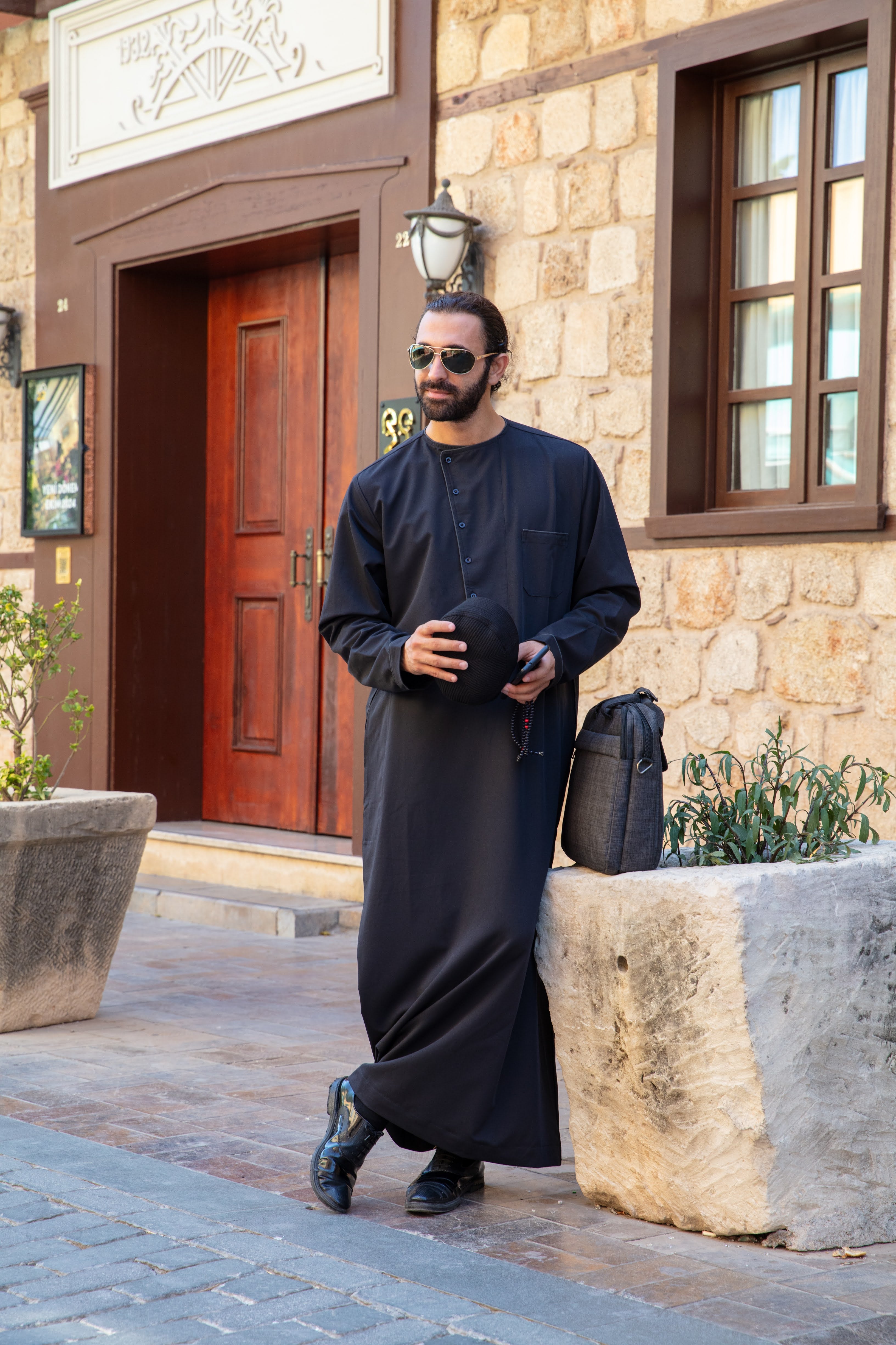 A person in a dark hooded robe, holding a string of beads, standing with back turned.