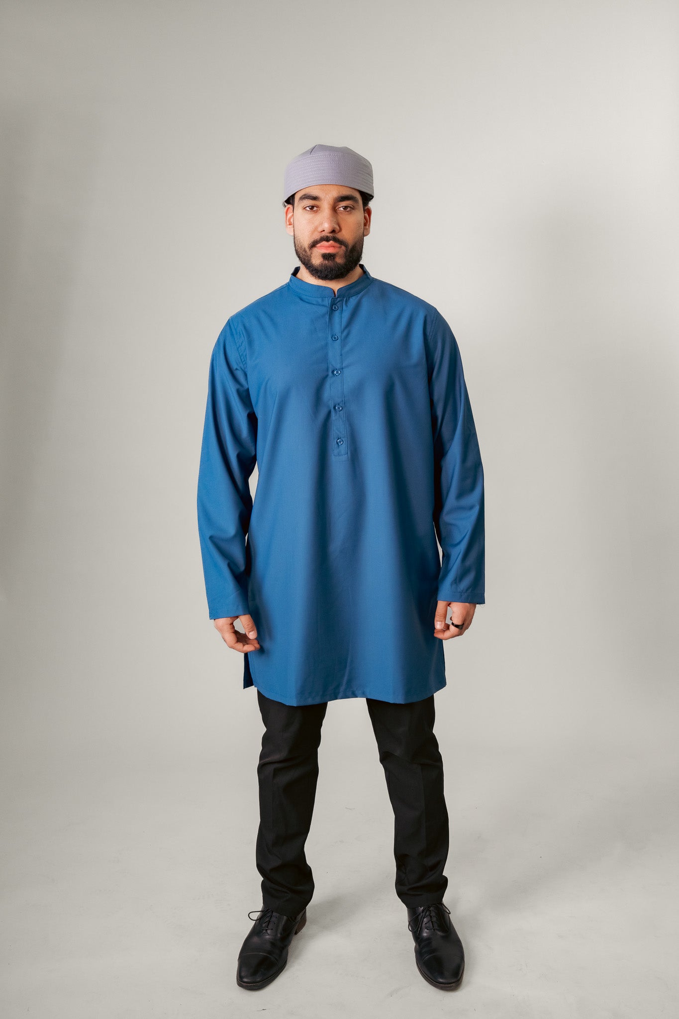 Man in navy traditional attire with hood, holding a beaded necklace, against a white background.