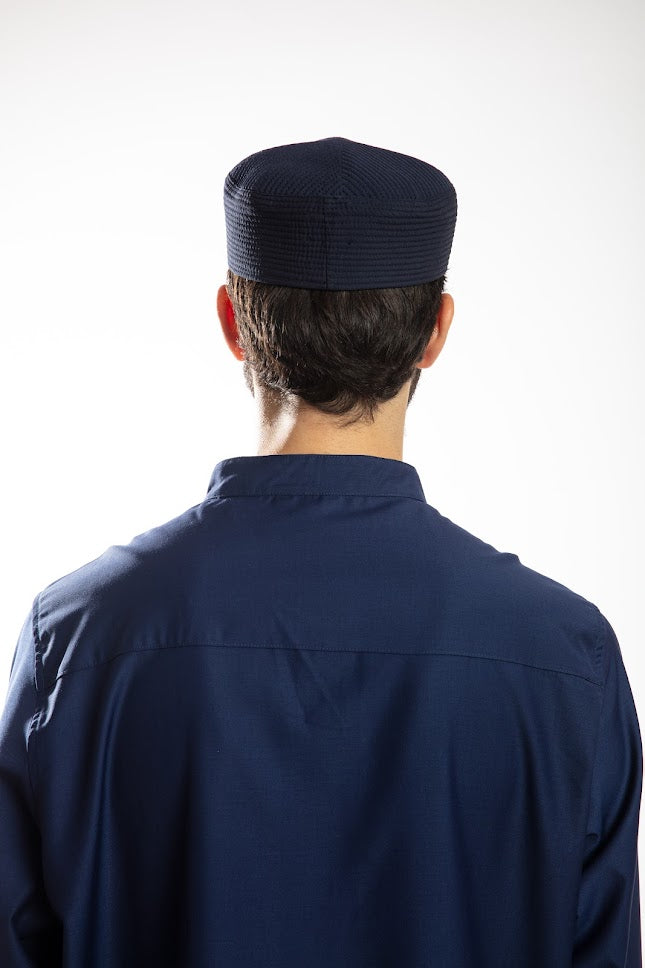 Man in navy traditional attire with hood, holding a beaded necklace, against a white background.