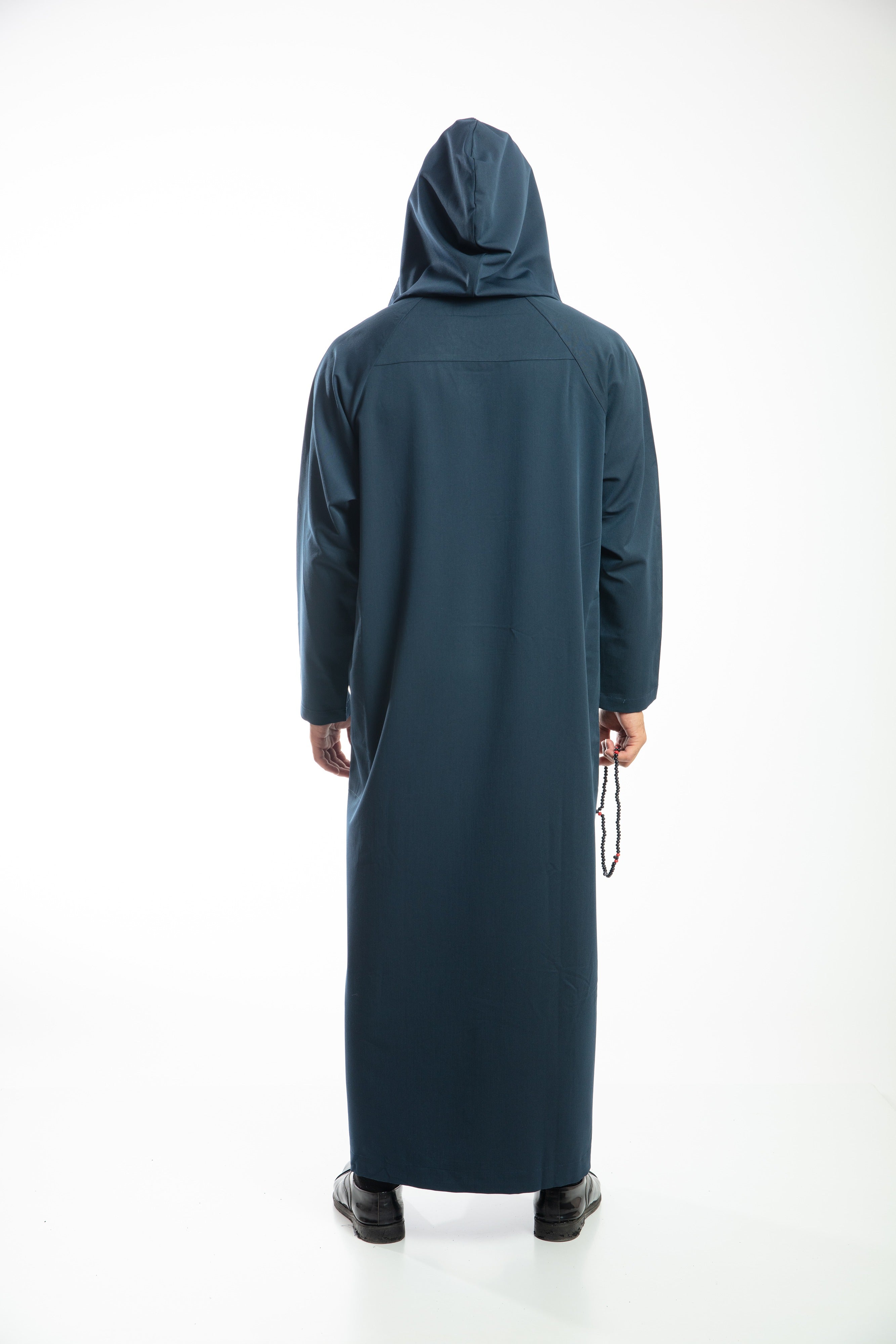 A person in a dark hooded robe, holding a string of beads, standing with back turned.