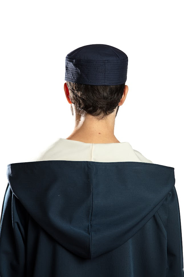 Man in navy traditional attire with hood, holding a beaded necklace, against a white background.