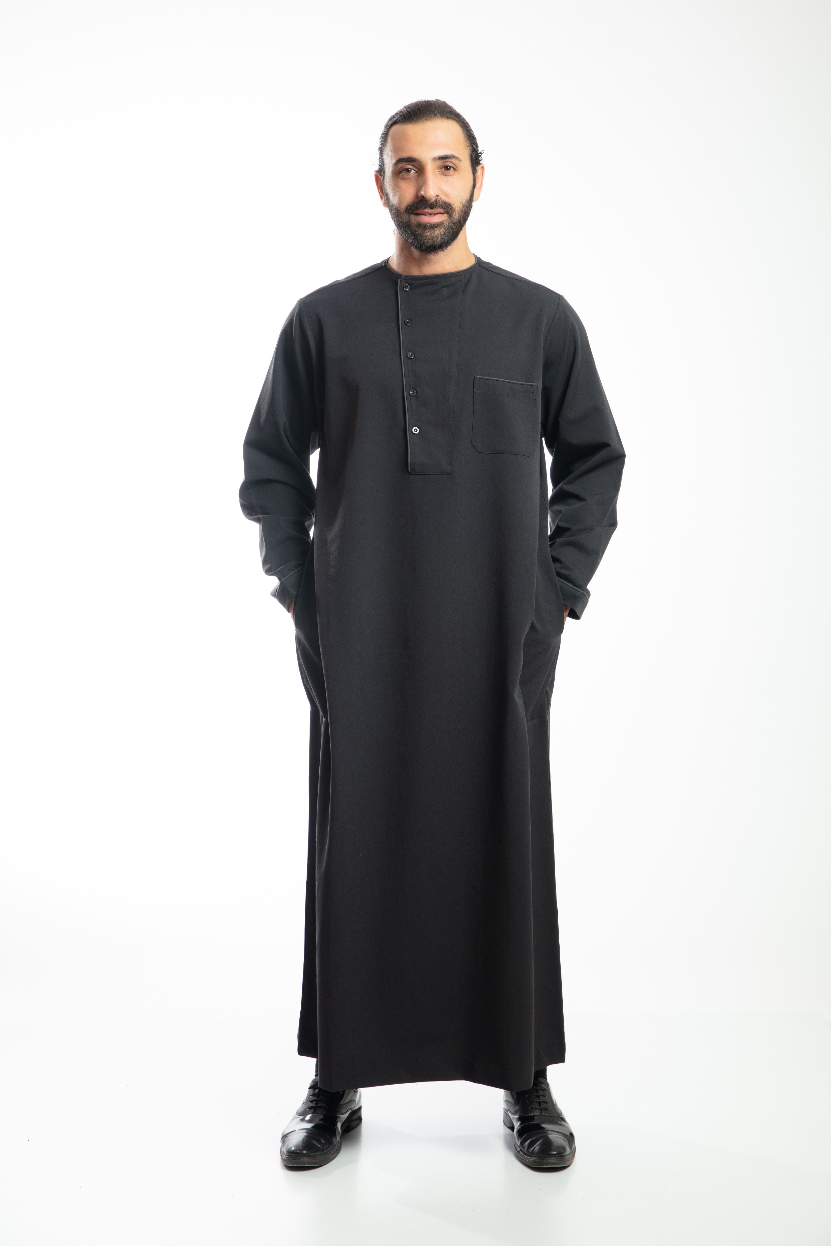 Man in navy traditional attire with hood, holding a beaded necklace, against a white background.